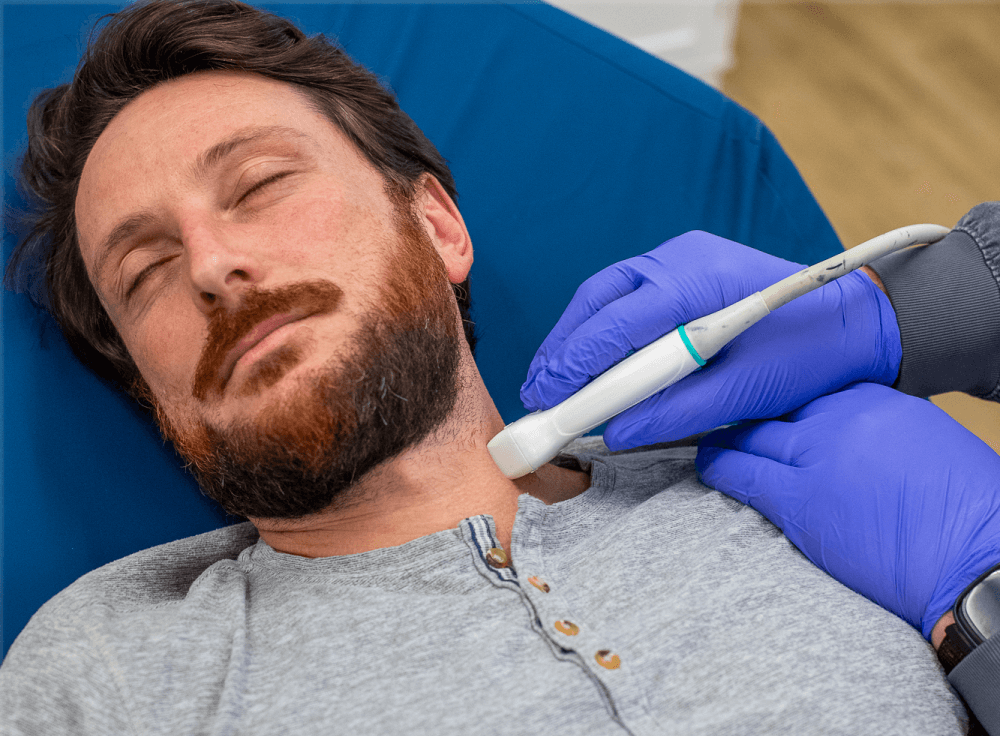 a man with facial hair laying back calmly with an ultrasound wand on his neck