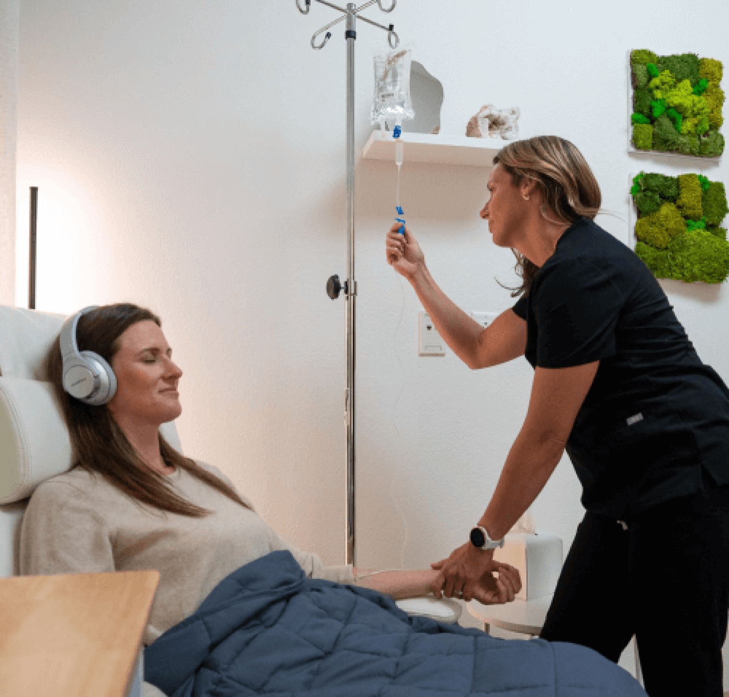 Photo of a woman monitoring an IV of a female patient wearing a blue weighted blanket and headphones in a white calm room with green moss art on the walls
